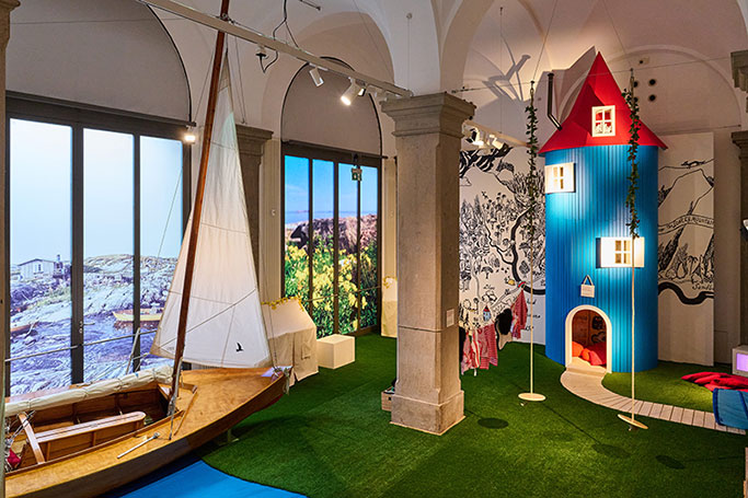 Raumansicht (Totale) der Tove Jansson-Ausstellung im Münchner Literaturhaus. Zu sehen ist ein historischer Saal mit Kunstrasen und einem symbolischen See. In ihm ist eine Spiellandschaft mit einem kleinen Segelboot und dem Muminhaus aufgebaut