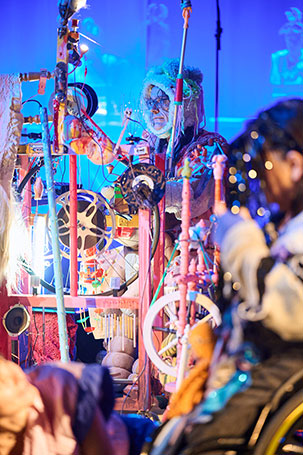 Aufnahme von Klaus Erika Dietl bei der Musikperformance Sharper Than A Needle in den Münchner Kammerspielen. Klaus sitzt hinter einem Aufbau aus bunten Gegenständen, die er zu einem Musikinstrument angeordnet hat und trägt ein Plüschtierkostüm