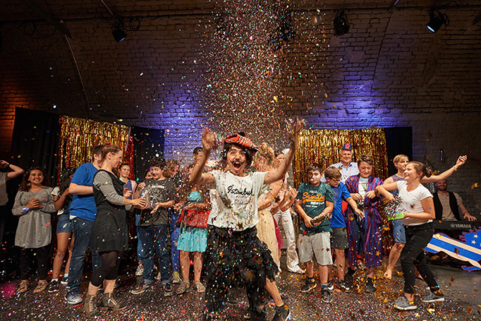 Aufführung des Vereins Impro macht Schule e.V. in München. Ein Kind steht lächelnd vor einer Gruppe weiterer Jugendlicher im Konfettiregen, der im Scheinwerferlicht glitzert. Theaterfotografie © Florian Freund